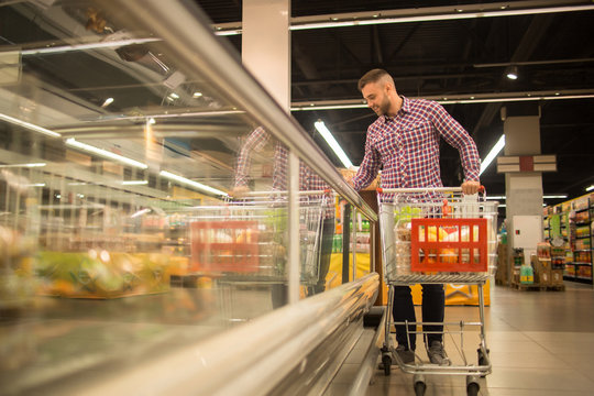 Full Length Portrait Of Handsome Young Man With Shopping Cart Choosing Groceries In Supermarket Looking At Frozen Foods In Freezer