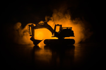 silhouette tracked excavator on dark foggy red orange sky background with toy car. Empty space