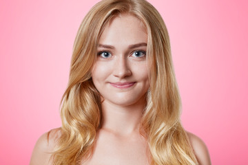 Naklejka premium Pleasant looking female with pure healthy skin, warm appealing eyes and charming smile, has long light hair, looks directly into camera, isolated over pink background. Beauty and youth concept