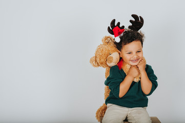Christmas portrait of adorable toddler boy wearing green pullover, reindeer ears with Santa Claus' hat, holding teddy bear