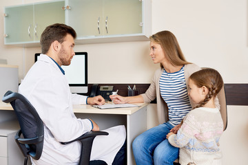 Fototapeta premium Portrait of little girl visiting doctor with mother, sitting at desk in office and consulting pediatrician for health check