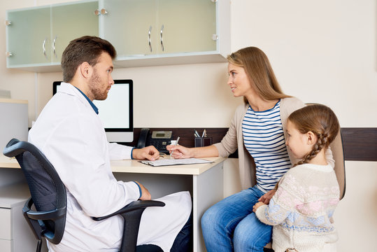 Portrait of little girl visiting doctor with mother, sitting at desk in office and consulting pediatrician for health check