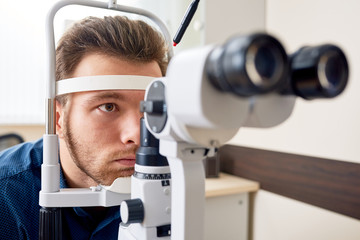 Portrait of young man  looking at slit lamp machine, resting head on stand during sight testing in optometrist office