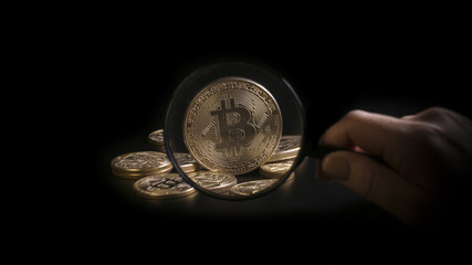 Unrecognizable person holding magnifying glass and looking through it on new virtual money golden bitcoin placed on a black background.