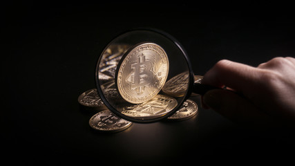 Fototapeta premium Unrecognizable person holding magnifying glass and looking through it on new virtual money golden bitcoin placed on a black background.