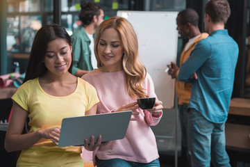 Interesting idea. Cheerful women are looking at laptop and smiling while leaning on table. Back view of involved men are standing around flipchart and discussing new business project in background