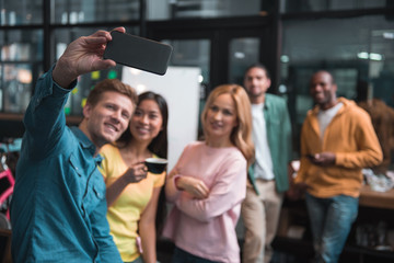 Friendly team. Selective focus of modern smartphone in hand of cheerful stylish guy. He is taking selfies with his qualified creative colleagues. They are standing in cozy office and smiling