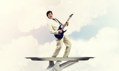 Businessman on metal tray playing electric guitar against blue sky background