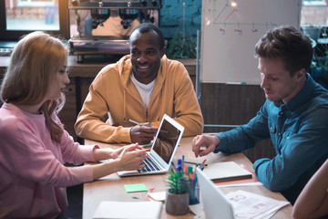 Portrait of cheerful man speaking with side view calm woman while locating at desk. She typing in laptop. Serious comrade situating opposite her. Work concept