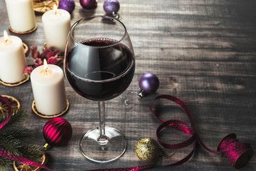 Glass of wine and burning candles. Red wine on wooden grey background. Slices  of dried orange. Christmas colorful baubles, fir-tree branches, ribbon