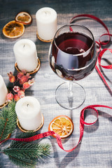 Glass of wine and burning candles. Red wine on wooden grey background. Slices  of dried orange. Christmas colorful baubles, fir-tree branches, ribbon