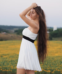 Fototapeta na wymiar Young brunette model at the white dress staying near the sunflowers field and looking forward