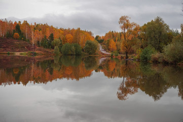 The autumn forest and the road on the hills are reflected in the calm water of the forest lake.