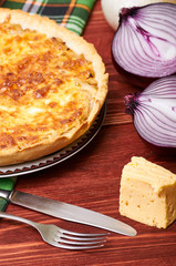 Appetizing French onion pie. A beautiful homemade pie with cheese crust stands on a green checkered napkin. Onions, cheese and cutlery on a rough wooden rustic table. Different kinds of onions.