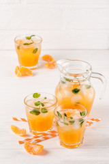 Mandarin cocktail with ice and mint in beautiful glasses and jug, fresh ripe citrus on white wooden background. Sweet orange juice. Close up photography. Selective focus