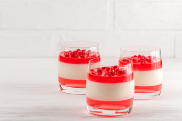 Creamy vanilla panna cotta with red jelly in beautiful glasses, fresh ripe pomegranate on white wooden background.