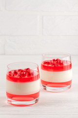 Creamy vanilla panna cotta with red jelly in beautiful glasses, fresh ripe pomegranate on white wooden background.