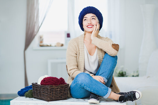 Beautiful woman in big blue knitted hat indoors sit on chest with basket yarn
