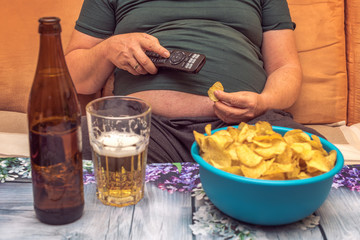 A fat man suffers from overweight and eats chips, drinks alcohol and looks in the telly. Concept:...