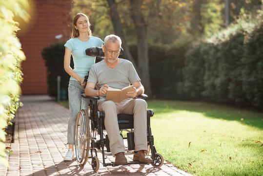 An elderly man is sitting in a wheelchair. Nearby is his daughter, she stands next to the old man's stroller.