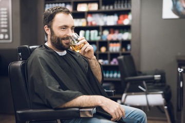 A man drinks alcohol in the hairdresser's armchair of a modern barbershop.