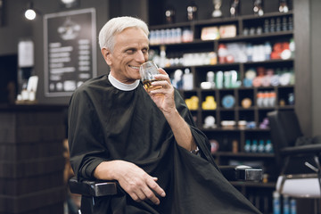 The old man drinks alcohol in the barber's chair in barbershop.