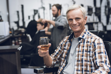 The old man drinks alcohol in the barber's chair in barbershop.