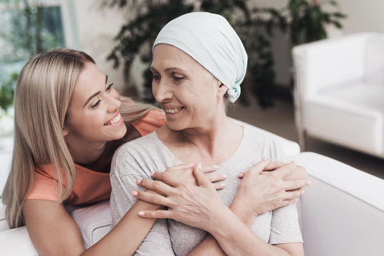 A woman with cancer is sitting on a white sofa next to her daughter. A girl is hugging a woman