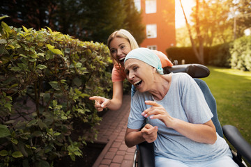 A woman with cancer is sitting in a wheelchair. She walks on the street with her daughter and they...