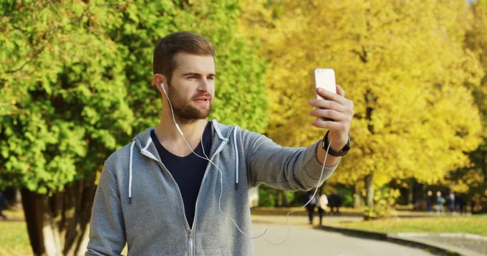 Young caucasian attractive sporty man in headphones having video chat on the smart phone in the sunny park in early autumn. Outdoors