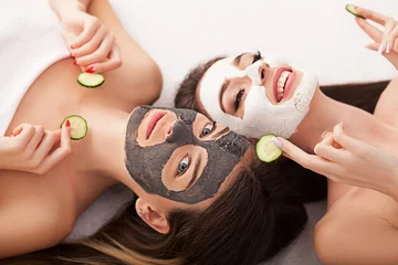 Tuinposter Home spa. Two women holding pieces of cucumber on their faces lying the bed. © Maksymiv Iurii