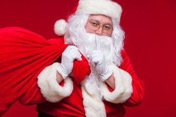 Portrait of Santa Claus with huge red sack keeping forefinger by his mouth and looking at camera