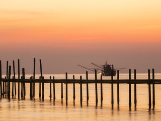 A wooden jetty is silhouetted at sunset on ko Kut island, Thailand. A shrimp fishing boat is in background.