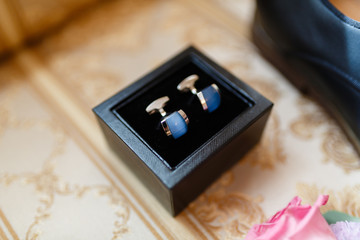 Cufflinks for the groom in the box on the background of the boutonniere with fresh flowers and classical leather shoes , men's accessories of a businessman on a sofa.