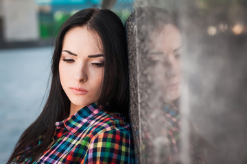 Fototapeta premium Portrait of young hipster girl with long hair leaning against the wall in the street, wearing colorful shirt in cage on the urban background