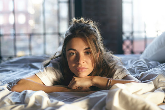 Horizontal shot of beautiful Caucasian lady with unbrushed hair lying on stomach on bed having bored look, thinking what to do during day, feeling lonely while her husband is away on business trip.