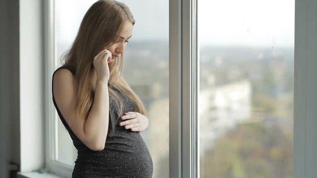 Smiling pregnant girl talking on her smartphone standing near window