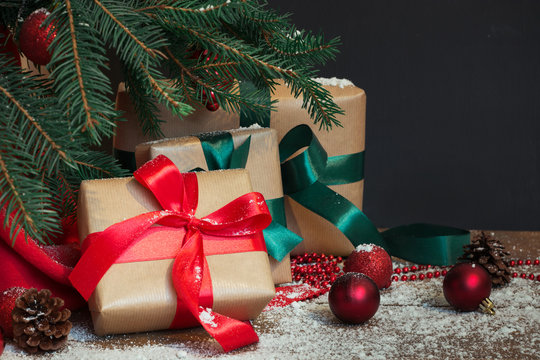 Christmas holiday background. Gifts with a red ribbon, Santa's hat and decor under a Christmas tree on a wooden board. Close up. Copy space on chalkboard.