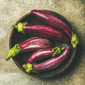 Flat-lay of fresh raw Fall harvest purple eggplants or aubergines in wooden bowl over concrete stone background, top view, square crop. Healthy Autumn vegan cooking ingredient