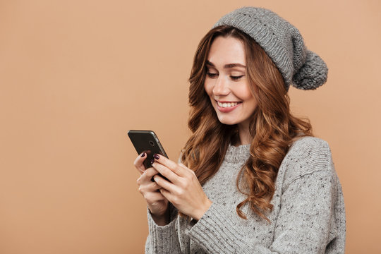 Cheerful brunette girl in  gray hat and knitted sweater using smartphone