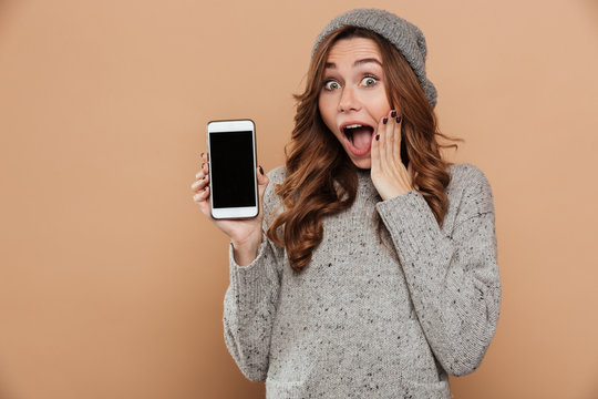 Close-up photo of amazed charming brunette woman in warm hat and knitted sweater showing blank mobile screen and looking at camera