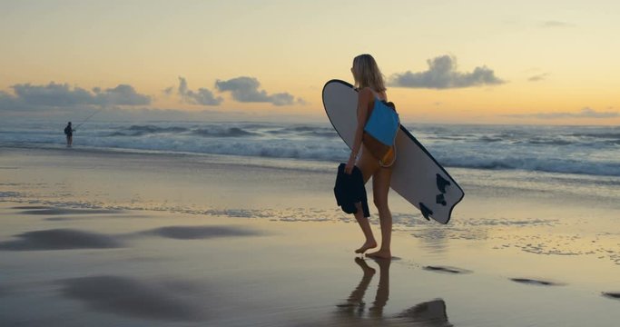 Beautiful Young Woman in the Swimsuit Walks Along the Beach while Carrying Surfboard. Sea with Waves and Sunset in the Background. Slow Motion.