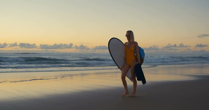 Beautiful Young Woman in the Swimsuit Walks Along the Beach while Carrying Surfboard. Sea with Waves and Sunset in the Background. Shot in Slow Motion.
