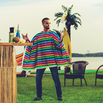 Handsome positive Mexican bartender in colorful poncho throws up a bottle of alcohol near the bar counter at the resort. Photo toned style Instagram filters