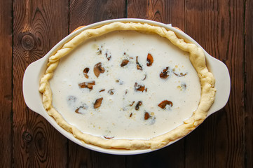 Half-finished quiche with mushrooms and cheese ready to bake in a ceramic dish on a wooden background.