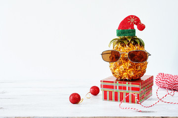 Closeup of pineapple in sunglasses and red hat with christmas decorations over white background. Copy space. Christmas in exotic country concept.