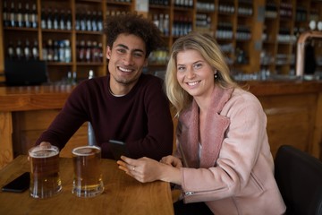 Smiling couple using mobile phone while having glass of beer in