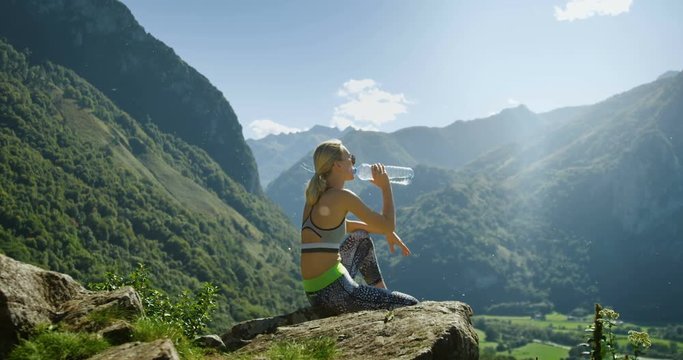 Fit Young Blonde Woman in Sportswear Sits on the Mountain and Drinks Water while Resting