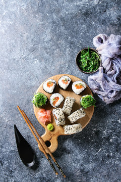 Homemade sushi rolls set with salmon, sesame seeds serving on wood serving board with pink pickled ginger, soy sauce, wasabi, seaweed salad, chopsticks on gray texture background. Top view, space