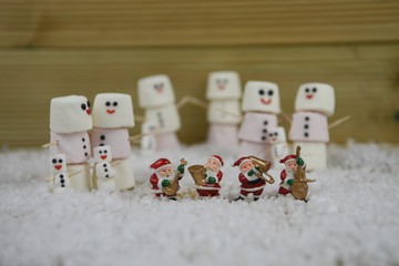 Christmas photography image using marshmallows shaped as snowman with iced on smile standing in snow with mini Santa Claus decorations and wood background